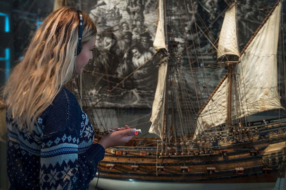 Kvinna med långt hår står och tittar på en monter med en stor modell av en segelskuta. Hon har hörlurar på sig och i handen håller hon en liten apparat, audioguiden. Bilden är tagen snett bakifrån.