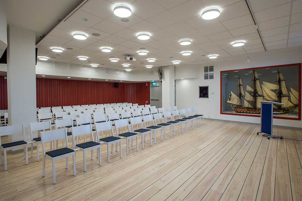 Sjöhistoriskas hörsal. Stolar uppställda i flera rader, vända mot ett talarpodium. Trägolv och tak med många lampor. Till höger i bilden en stor glasmonter med en modell av ett segelfartyg.
