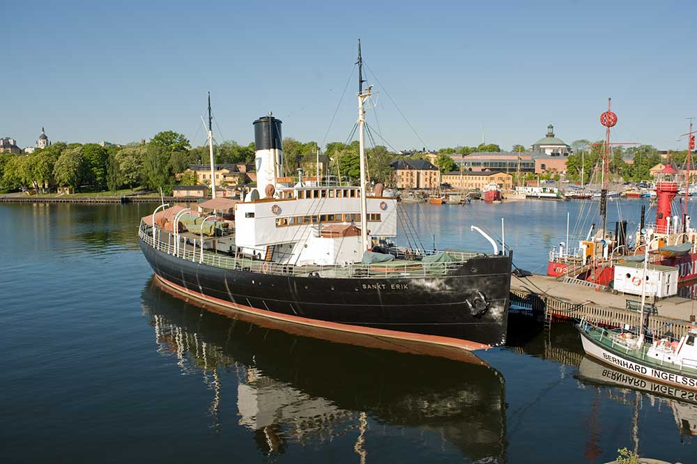 Isbrytaren Sankt Erik ligger vid museifartygspiren. På andra sidan piren ligger fyrskeppet Finngrundet. Vattnet är lugnt, nästan spegelblank. I bakgrunden syns Skeppsholmen. På andra sidan