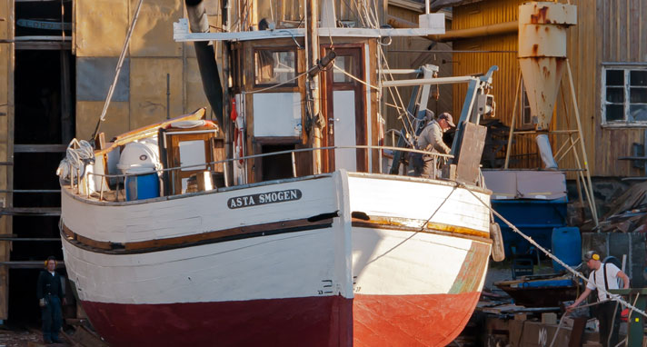 Fiskebåten Asta av Smögen ligger på slipen till ett varv och två män håller på med några tampar på barbordsida i solskenet. Hon syns akterifrån med styrhytten och en del fast utrustning för fiske syns uppe på däck. Skrovet är vitmålat.  