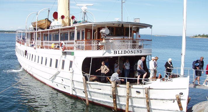 Passagerarångbåten Blidösund är en stor vitmålad skärgårdsbåt med två salongsdäck för passagerare. Den övre våningen är indragen så att det finns ett promenaddäck runtom. På taket syns en livbåt i trä och en ljusgul skorsten. I fören håller passagerarna på att stiga av.
