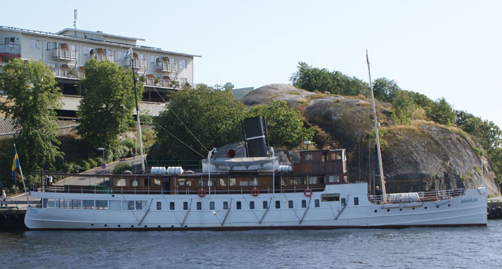 Den stora passagerarångbåten Bohuslän ligger förtöjd vid en klippa och visar upp sin styrbordssida. Hon har två salongsvåningar med ett promenaddäck upptill. Skrovet är vitmålat medan övre däcket och styrhytten är brunt trä-färgade. Hon har två master och bakom styrhytten en svart skorsten midskepps.