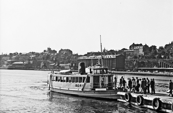 Den lilla passagerarbåten Bore ligger med fören mot en brygga invid Slussen i Stockholm. På det svartvita fotot står passagerarna redo att kliva ombord på fartyget. Fartyget har bara en våning och i fören en uppstickande styrhytt. Midskepps syns en låg svart skorsten och längst bak en uppochnedvänd livbåt.