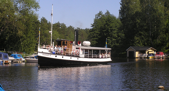 Boxholm två är ett mindre passagerarfartyg med svart skrov med vitmålad överdel. På fördäck syns passagerare och akter om den bruna styrhytten finns ett taktäckt öppet däck. Fartyget syns under gång på en sjö med grönska och småbåtar i bakgrunden.  