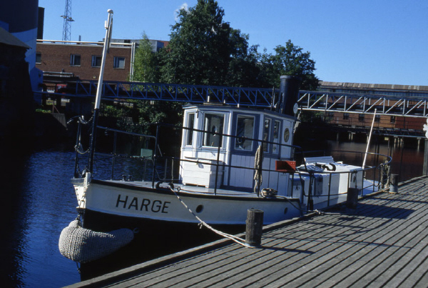 Harge är en mindre vitmålad bogserbåt med svart skrov. Styrhytten står midskepps och längs relingen finns ett svart mantåg för att hålla passagerarna säkra. På bilden ligger Harge vid kaj i hamnen i Karlstad. 
