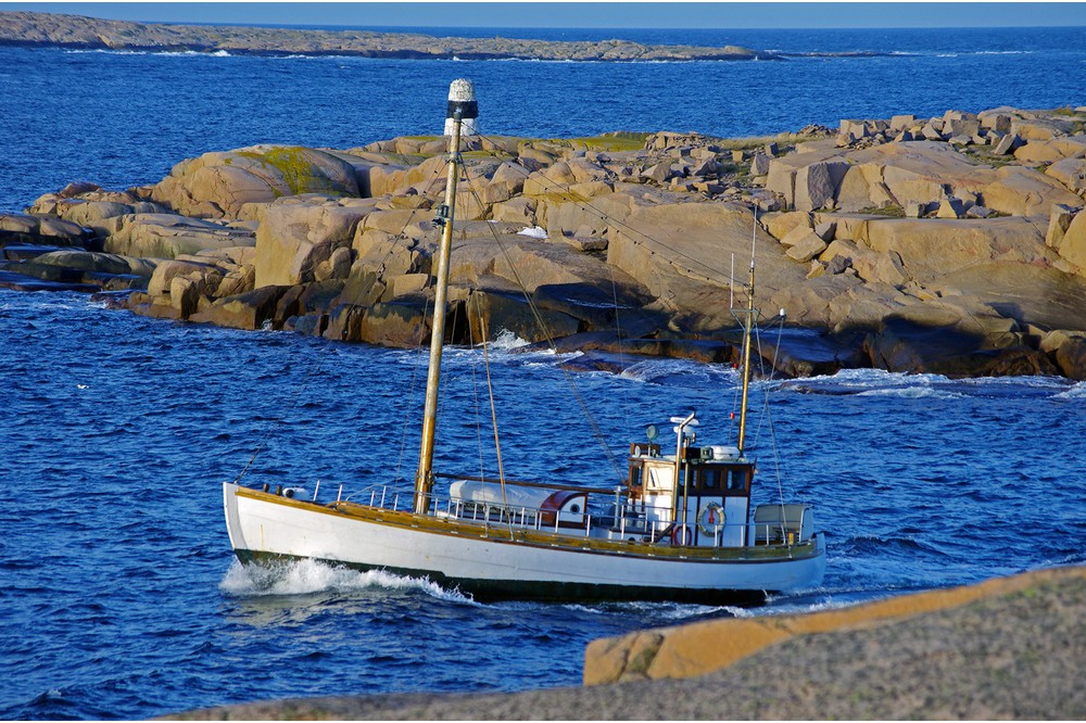 Heimdal är en lotsbåt med vitt träskrov, mast och styrhytt i aktern. På bilden är fartyget under gång med bohuslänska klippor i bakgrunden. 