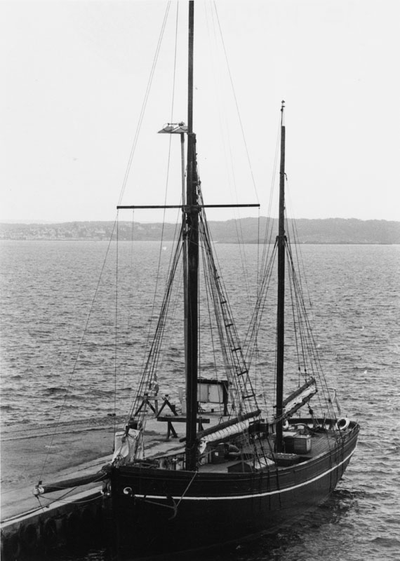 Lord Nelson är ett tvåmastat segelfartyg med seglen i sina kapell vid bommen. Däcket ser prydligt städat ut och hon har ett svart skrov. På den svartvita bilden ligger fartyget förtöjt vid en pir. 