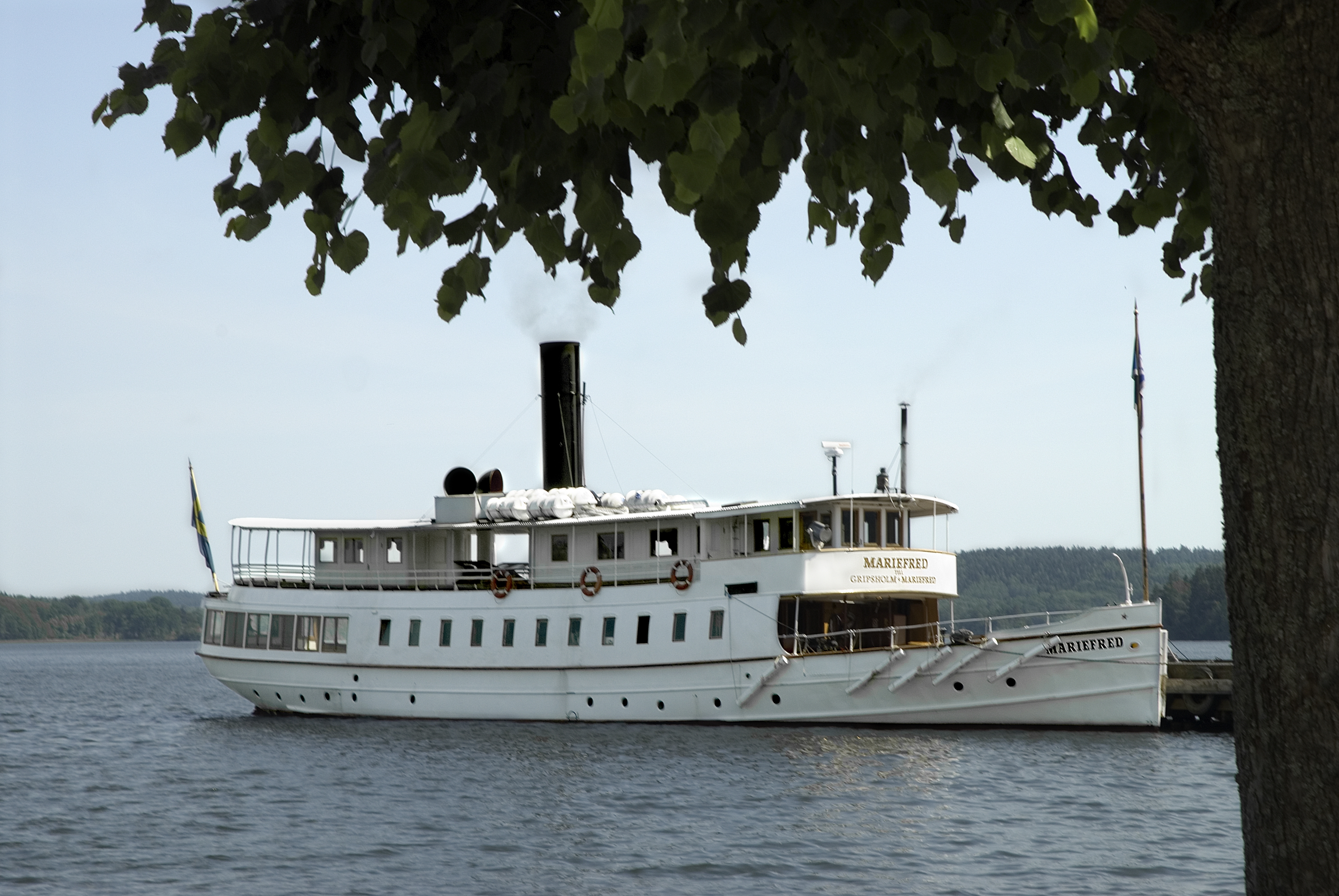 Mariefred är ett vitmålat passagerarångfartyg med salonger på två däck. Ur den svarta höga skorstenen stiger ljusgrå rök. På bilden ligger fartyget förtöjt vid en brygga i Mälaren. 