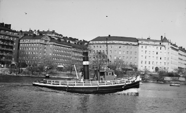 Örnen är en bogserbåt med svart skrov med vit reling och mantåg. Styrhytten ligger föröver och midskepps syns en skorsten. På den svartvita bilden är fartyget ute och kör på Liljeholmsviken med Bergsunds strand i Stockholm i bakgrunden. 