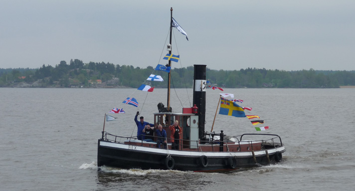 Rex är en liten ångbogserbåt med styrhytt och skorsten midskepps.  Skrovet är svartmålat med en vit rand upp mot relingen. I masten finns ett flaggspel av nationsflaggor hissat. På bilden är fartyget ute och kör i Mälaren och på fördäck står besättningen och vinkar. 