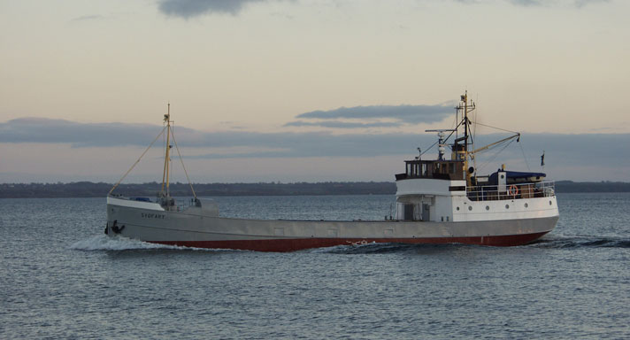 Olof Trätälja är ett traditionellt lastfartyg med gråmålat skrov och vit överbyggnad i aktern. På bilden är fartyget ute till sjöss. 