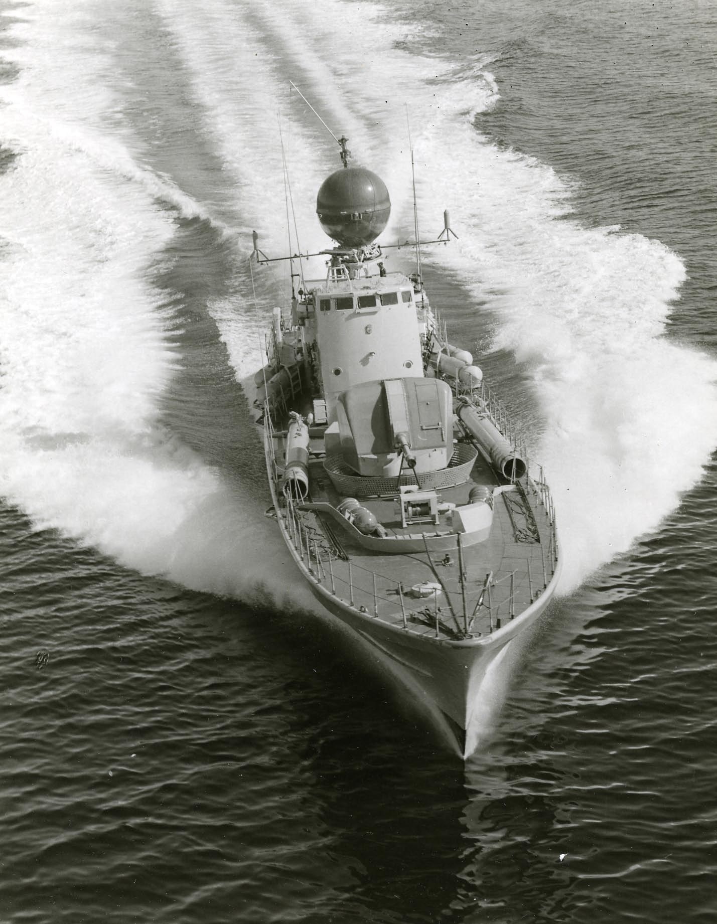 Militärfartyget Spica är en torpedbåt som går i full fart så att svallvågorna yr. På däck syns en automatkanon, bakom den en högre kommandobrygga och ovanpå den radar skyddad av ett klot. Torpedtuber syns på bägge sidor om kanonen. 