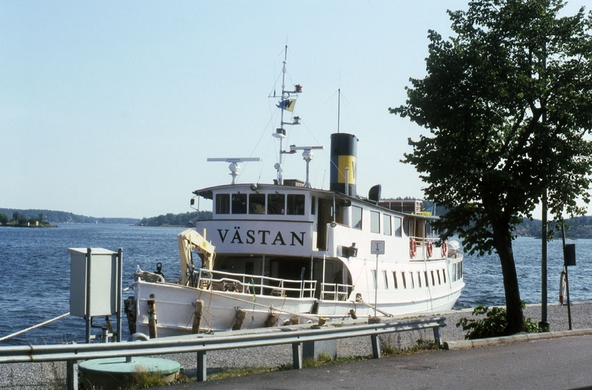 Västan är ett vitmålat passagerarfartyg med två däck. På fördäck står en gul kran och föröver på övre däck syns en kommandobrygga. På skorstenen syns Waxholmsbolagets rederimärke i gult och blått. På bilden ligger fartyget vid kaj i Stockholms skärgård. 